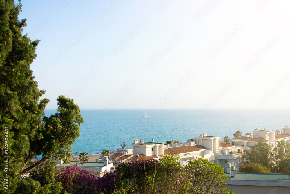 A view to Mediterrain sea from a view point over the tile roofs of white houses and hotels, green trees and pink violet colorful bougainvillea flowers at the foreground.