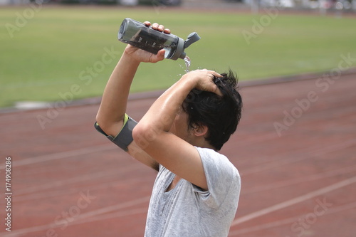 Healthy young Asian runner pouring water with waterbottle on his face after running on track in stadium.