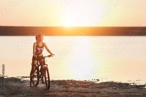 A strong blonde woman in a colorful suit sits on the bicycle near the water at sunset on a warm summer day. Fitness concept. Sky background