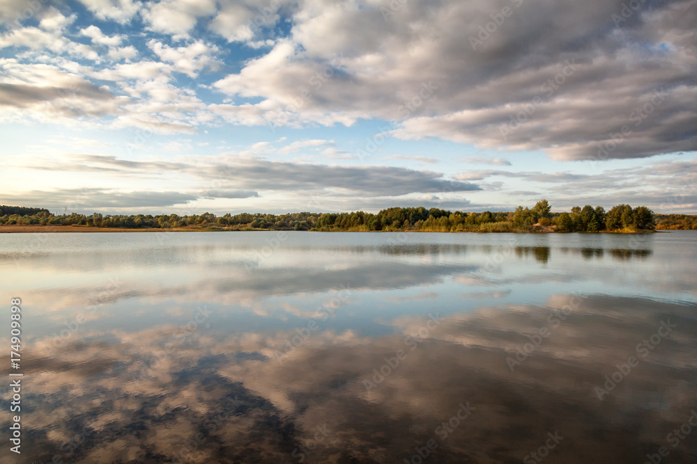 A beautiful autumn landscape, a forest reflected in a river, the sky with clouds.