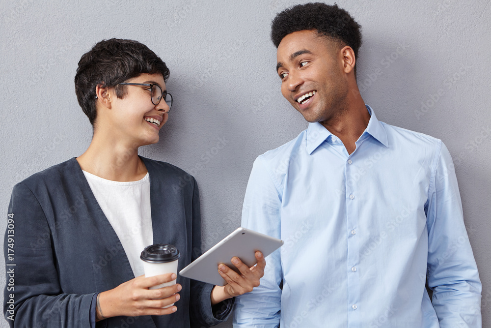 Stylish mixed race woman with short haircut holding digital tablet and papercup with takeaway coffee and talking to handsome African American man while standing in queue for doctor or job interview Stylish mixed race woman with short haircut holding digital tablet and papercup with takeaway coffee and talking to handsome African American man while standing in queue for doctor or job interview