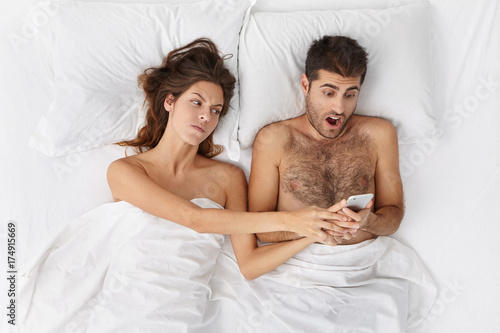 Unhappy shocked bearded husband caught his wife cheating on him, holding mobile phone and reading text messages from her lover, angry woman trying to take away gadget from her husband's hands photo