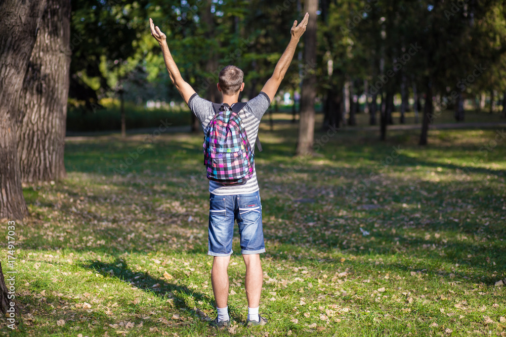 Young guy standing in park with backpack and hands up at sunset back view. Traveler, hipster standing in the woods, Hiking, Forest, Journey.