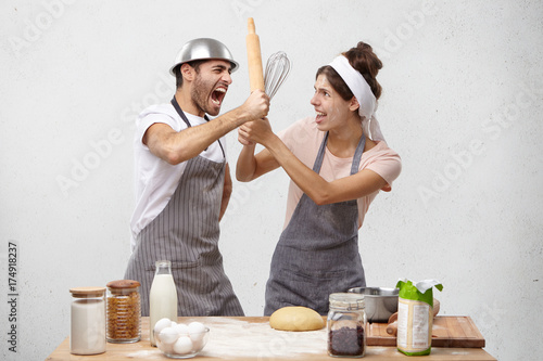 Furious outraged man finds out about wife`s betrayal, fight with kitchen untensils, screams in bewilderment and panic. Cook competitiors have battle at work place, defend by rolling pin and whisk photo