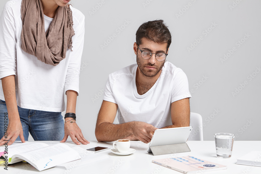 Working in team. Candid shot of handsome young bearded chief engineer checking project of his unrecognizable female employee, sitting at desk and pointing at digital tablet, explaining mistakes Working in team. Candid shot of handsome young bearded chief engineer checking project of his unrecognizable female employee, sitting at desk and pointing at digital tablet, explaining mistakes