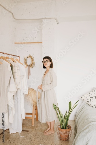 Attractive brunette young woman wears glasses and linen beige robe at home standing in her white room in front of rail with clothes in morning.