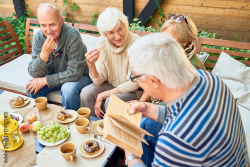 Group of joyful senior friends having fun outdoors: they enjoying delicious pastry and fragrant coffee while elderly man in striped sweater reading their favorite novel aloud