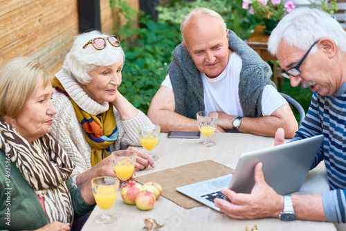 Two senior couples spending weekend together at backyard: they looking through photos on laptop and enjoying freshly squeezed juice
