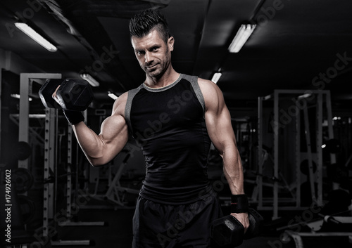 Portrait of sporty healthy strong muscle charismatic happy smiling handsome man bodybuilder hard training workout in well-equipped gym doing biceps exercises with dumbbells in black t-shirt outfit