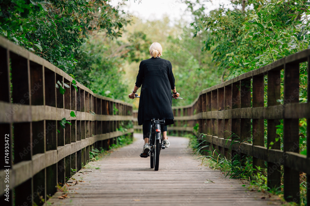 Young stylish blonde woman in black cardigan riding on bicycle on wooden road in park. Handsome girl having a ride. Back view Young stylish blonde woman in black cardigan riding on bicycle on wooden road in park. Handsome girl having a ride. Back view