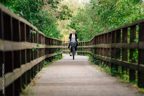 Young stylish blonde woman in black cardigan riding on bicycle on wooden road in park. Handsome girl having a ride.