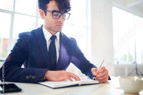 Young confident manager in formalwear writing plan of his working day in office