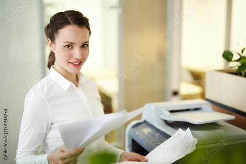 Quadro em tela Young secretary with papers standing by photocopier in office