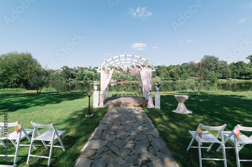 Beautiful summer wedding ceremony outdoors. Decorated chairs stand on the grass. Wedding arch made of light cloth and white and pink flowers on a green natural background. Wooden doors. Empty seats