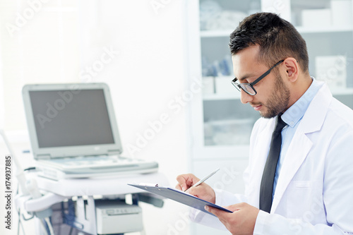 Profile view of handsome young physician in eyeglasses filling in medical card while consulting patient at office, portrait shot