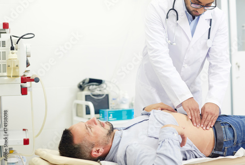 Concentrated young physician doing abdominal palpation of middle-aged patient suffering from pain, interior of modern office on background photo