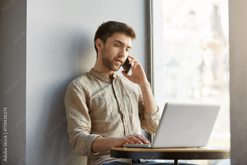 Unhappy handsome guy with dark hair, sitting in cafe, working on laptop computer and talking with dissatisfied customer on phone. Unhappy handsome guy with dark hair, sitting in cafe, working on laptop computer and talking with dissatisfied customer on phone.