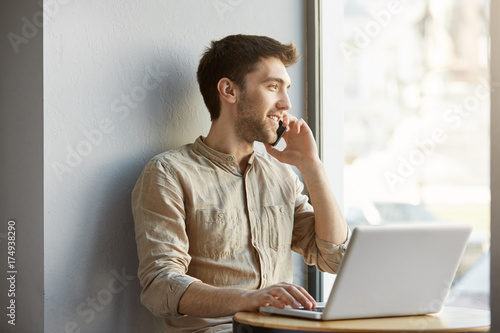 Joyful attractive shaved guy sitting in coworking space, working on laptop computer and talking on phone with girlfriend about date in evening.