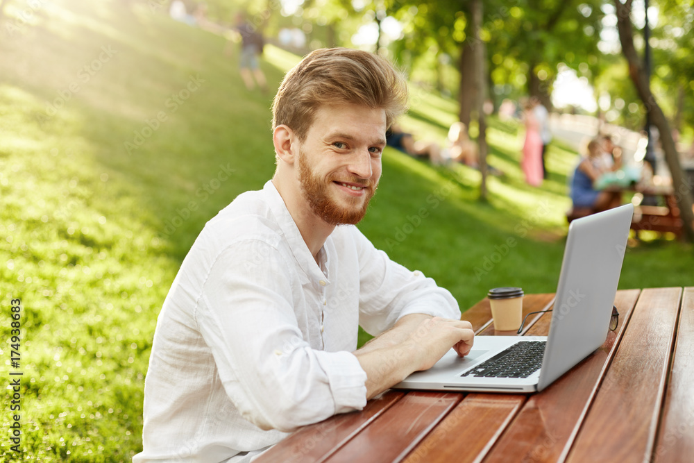 Mature ginger handsome man with beard in casual stylish clothes smiles, sitting in park, looking at camera posing with laptop for article about freelance. Mature ginger handsome man with beard in casual stylish clothes smiles, sitting in park, looking at camera posing with laptop for article about freelance.