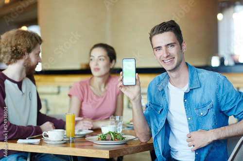 Happy guy with smartphone showing promo advert of new diner where he having lunch with his friends
