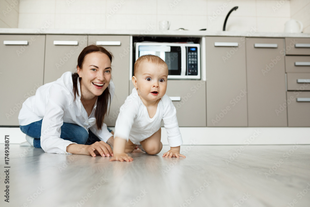 Young handsome mother standing on knees in kitchen playing with her lovely newborn son. Child happily crawls in camera direction. Young handsome mother standing on knees in kitchen playing with her lovely newborn son. Child happily crawls in camera direction.