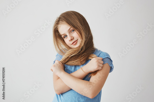 Beautiful joyful light-haired girl in blue shirt, looking in camera with funny face expression, holding hair in hands and crossing it.