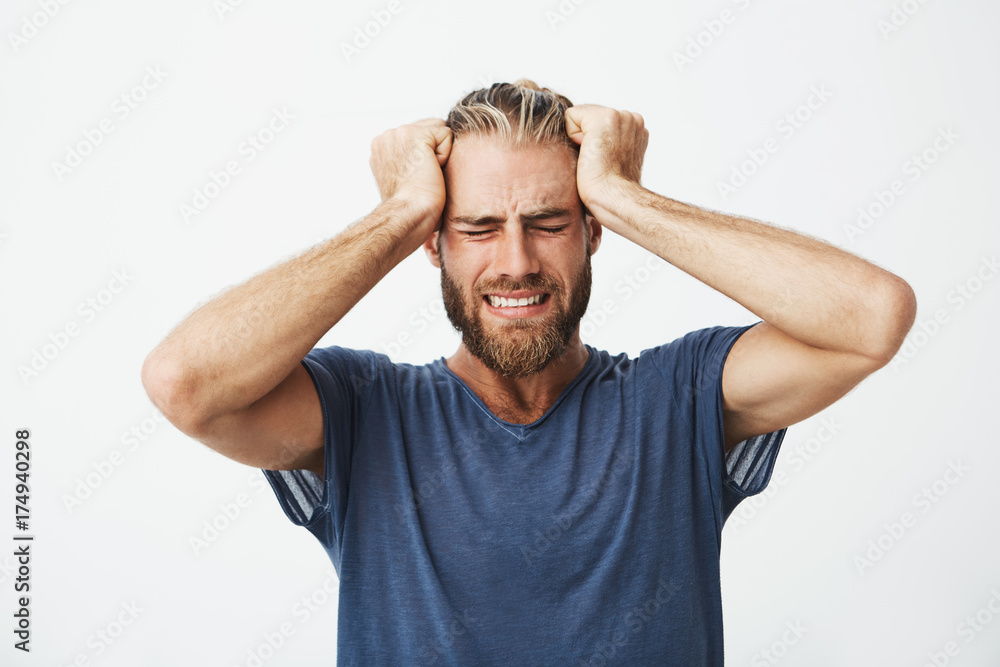 Portrait of beautiful nordic guy with trendy haircut and beard holding head with hands suffering from headache after a long day at work.