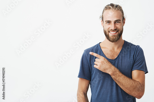 Happy beautiful bearded guy with good-looking hairstyle looking at camera, smiling and pointing aside with hand. Copy space.