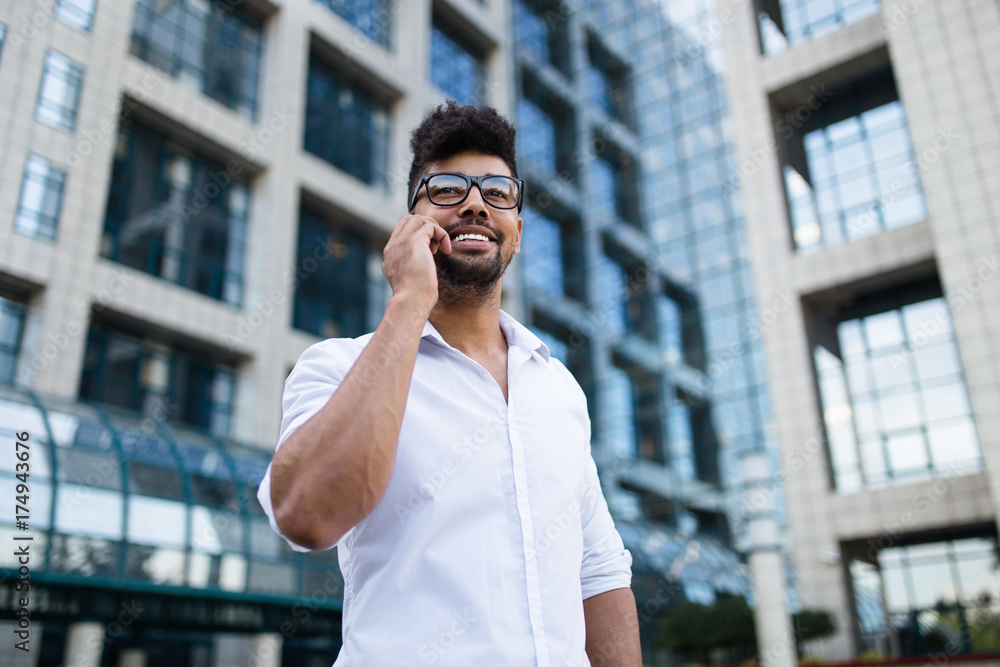 Positive and handsome Afro American man standing in front of huge modern business building smiling and talking on cell phone Positive and handsome Afro American man standing in front of huge modern business building smiling and talking on cell phone