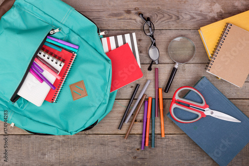 Backpack and school supplies: books, magnifying glass, notepad, felt-tip pens, eyeglasses, scissors on brown wooden table