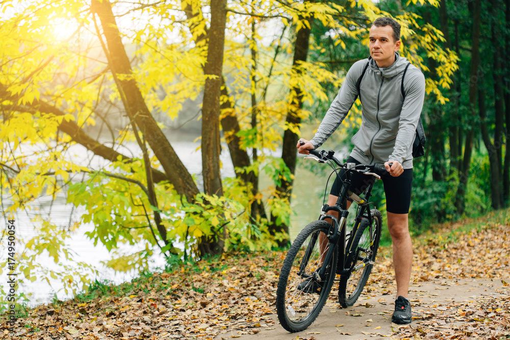 Young pretty athletic man standing with bicycle in autumn park. Fall season background. Male cyclist in forest