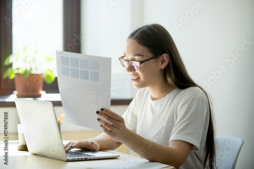 Young happy designer woman working on laptop holding pantone color swatch palette for house interior renovation project. Attractive model choosing color samples, doing home design using pc software