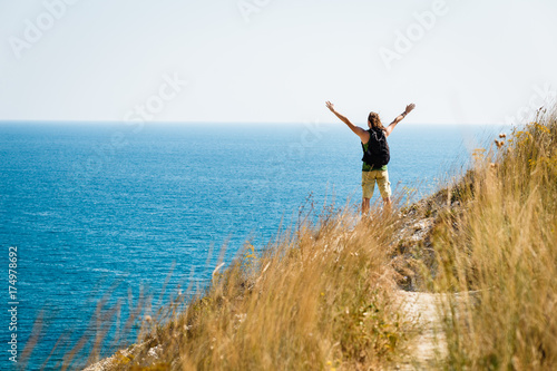 Young man traveler with backpack relaxing on cape near the cliff him arms open felling freedom and enjoying a holiday on the sea shore at sunny day. Adventure, travel people concept. Vacation.