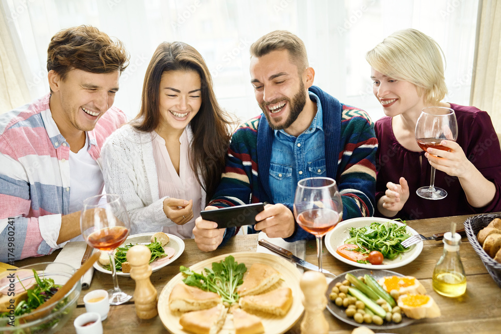 Portrait of four happy people laughing video chatting from smartphone at dinner table during holiday celebration
