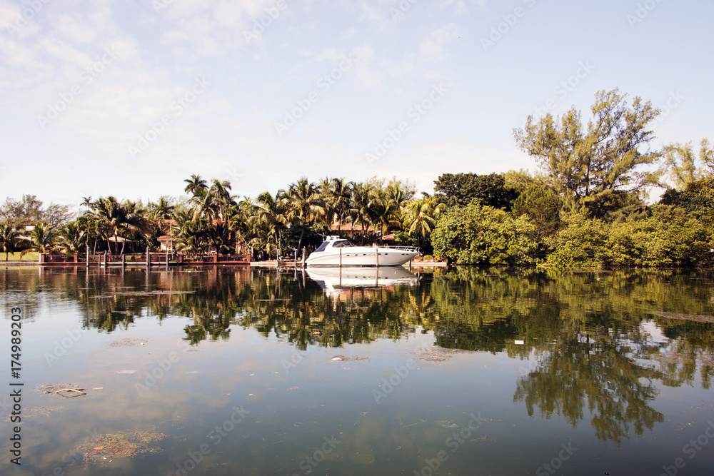 Fototapeta premium A view and a mirror image reflection of a motorboat, house villa and trees and landscape on the river in Miami Beach, Florida, USA