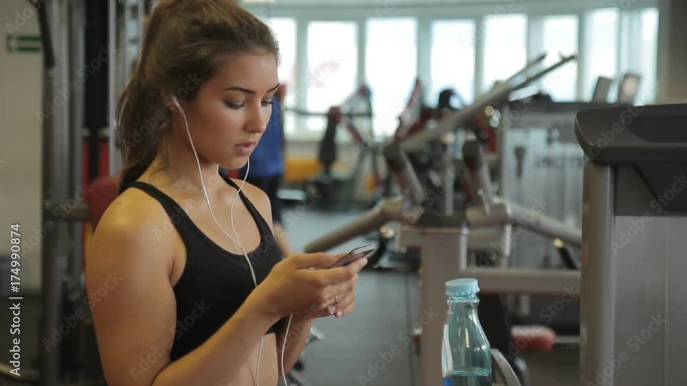 Young woman in headphones with phone standing in gym indoors. She looks attentively at screen of smartphone and types message in between exercises. Young woman in headphones with phone standing in gym indoors. She looks attentively at screen of smartphone and types message in between exercises.