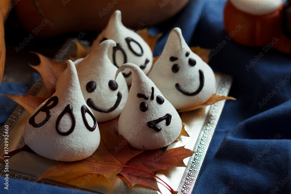 Funny meringue ghosts for Halloween party on autumn decorations. Golden leaves, pumpkin, blue textil and light of candles. Halloween concept