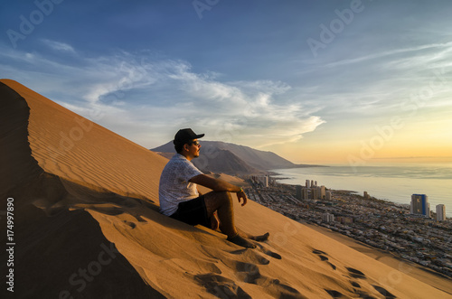 Young man sitting at the summit of enormours sand dune and overlooking city and ocean at the sunset