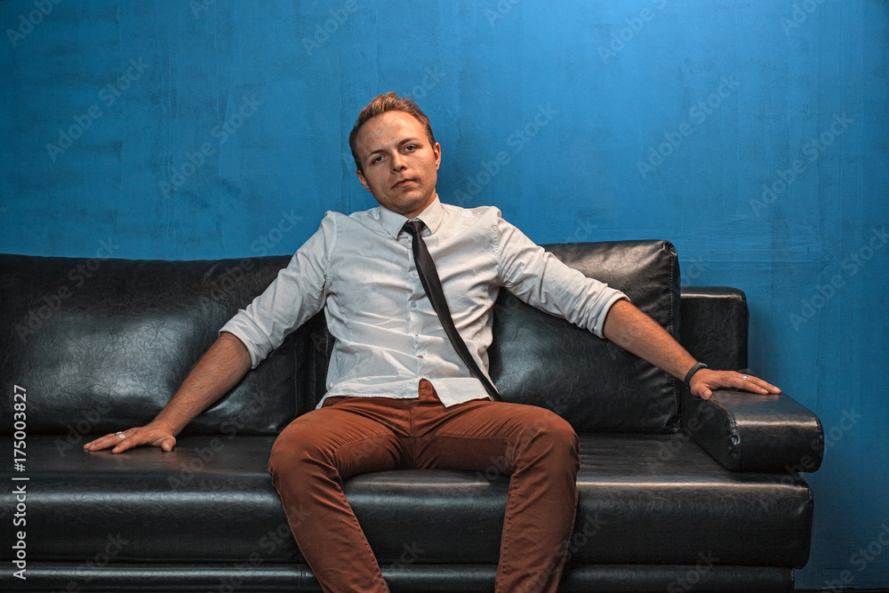 A handsome young man with modern haircut is sitting on a black leather couch in the studio. A handsome young man with modern haircut is sitting on a black leather couch in the studio.