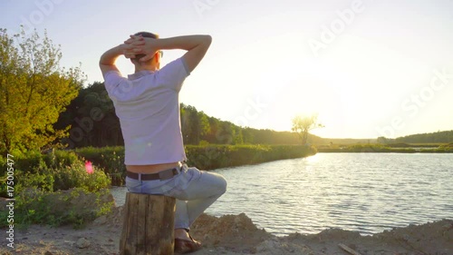 Relaxing young man sits on a stump on the lake, with his hands behind his head