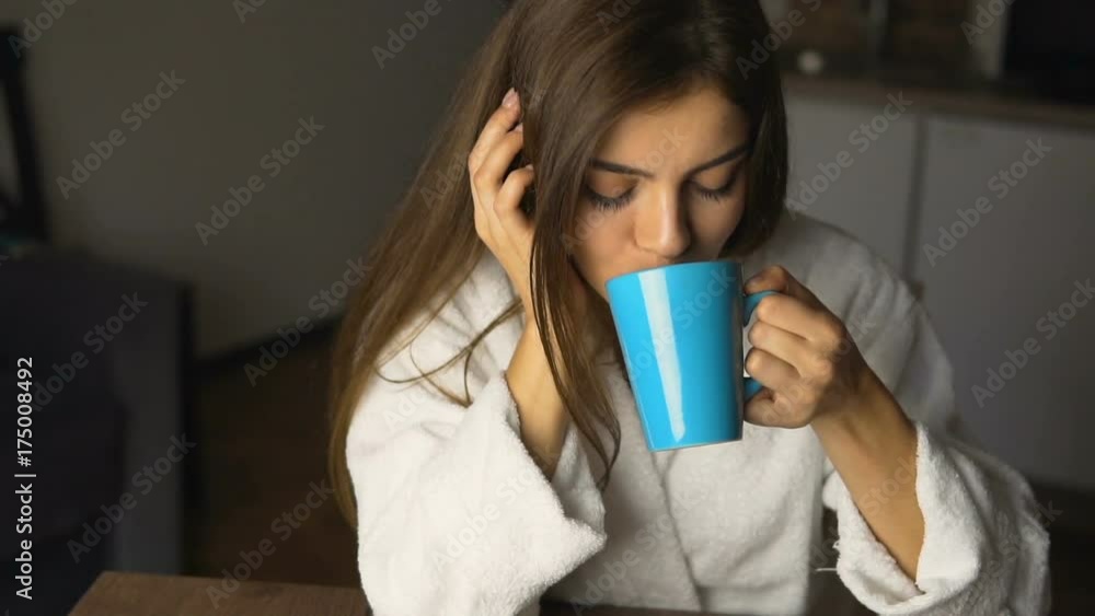 Beautiful caucasian girl, in a white bathrobe, enjoys morning coffee time in the kitchen, slow motion Beautiful caucasian girl, in a white bathrobe, enjoys morning coffee time in the kitchen, slow motion
