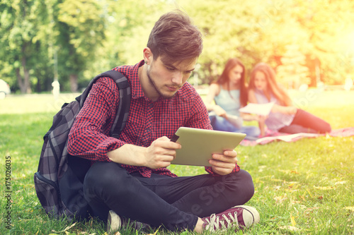 Student life. Young male student reads your electronic digital tablet sitting on the green lawn near the university.