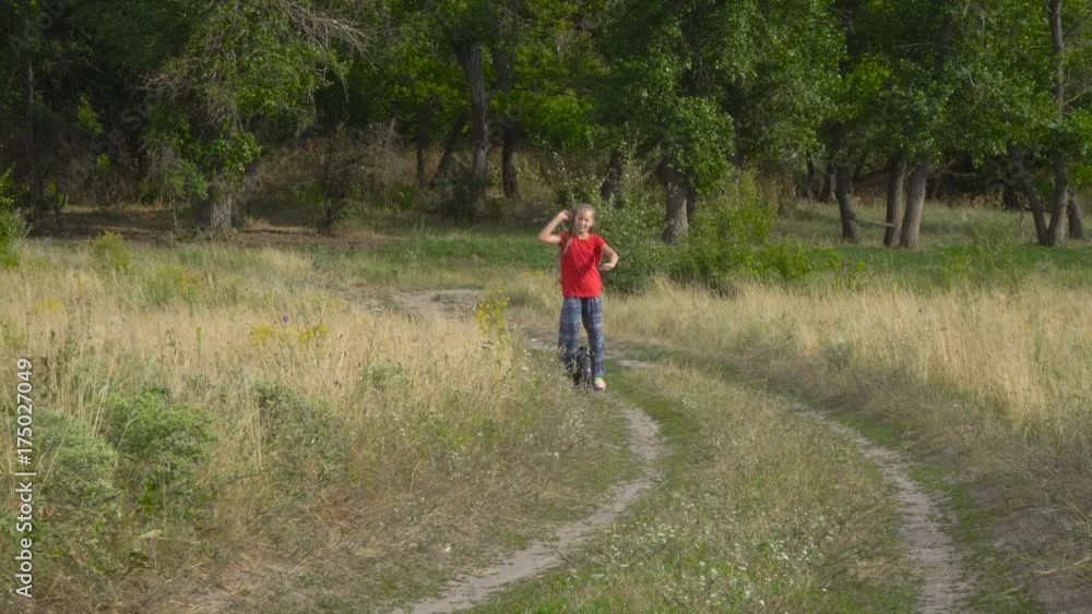 The girl is riding on a monocycle along the field road. The child is wearing a yellow jersey. She is happy and smiling. The girl is riding on a monocycle along the field road. The child is wearing a yellow jersey. She is happy and smiling.