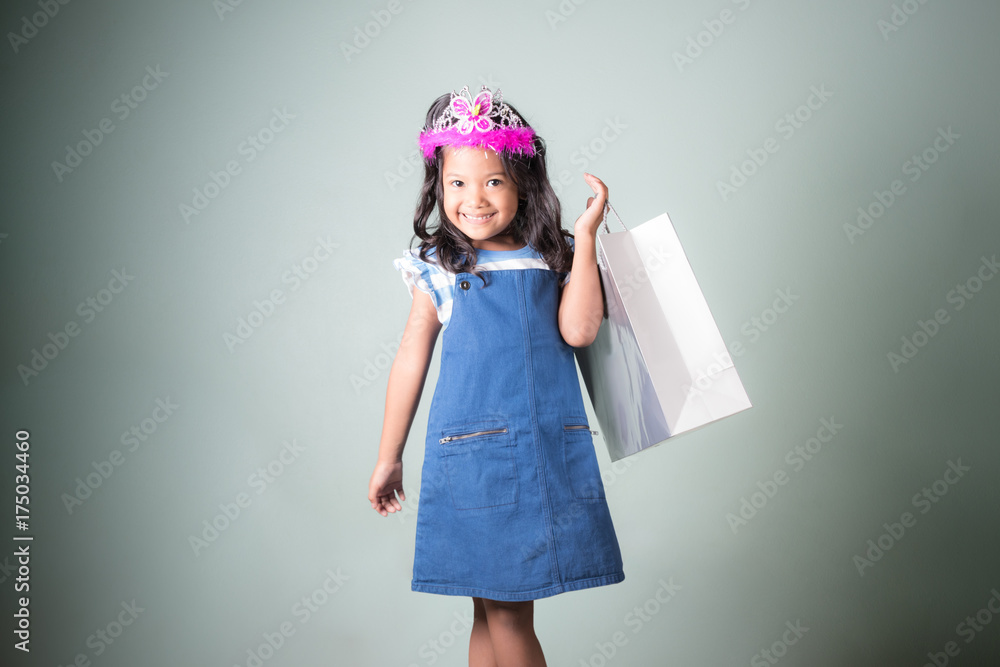 pretty little asian girl in blue jeans dungarees holding shopping bags with white concrete wall background.portrait of young kid smiling and looking at camera pretty little asian girl in blue jeans dungarees holding shopping bags with white concrete wall background.portrait of young kid smiling and looking at camera