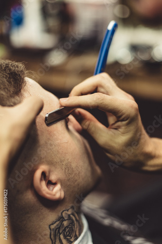 Anonymous barber shaving stylish tattooed man with razor in barbershop.