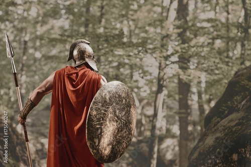 Back view of man in gladiator helmet and red long cloak standing with round shield and spear in hands looking away in woods. Spartan.
