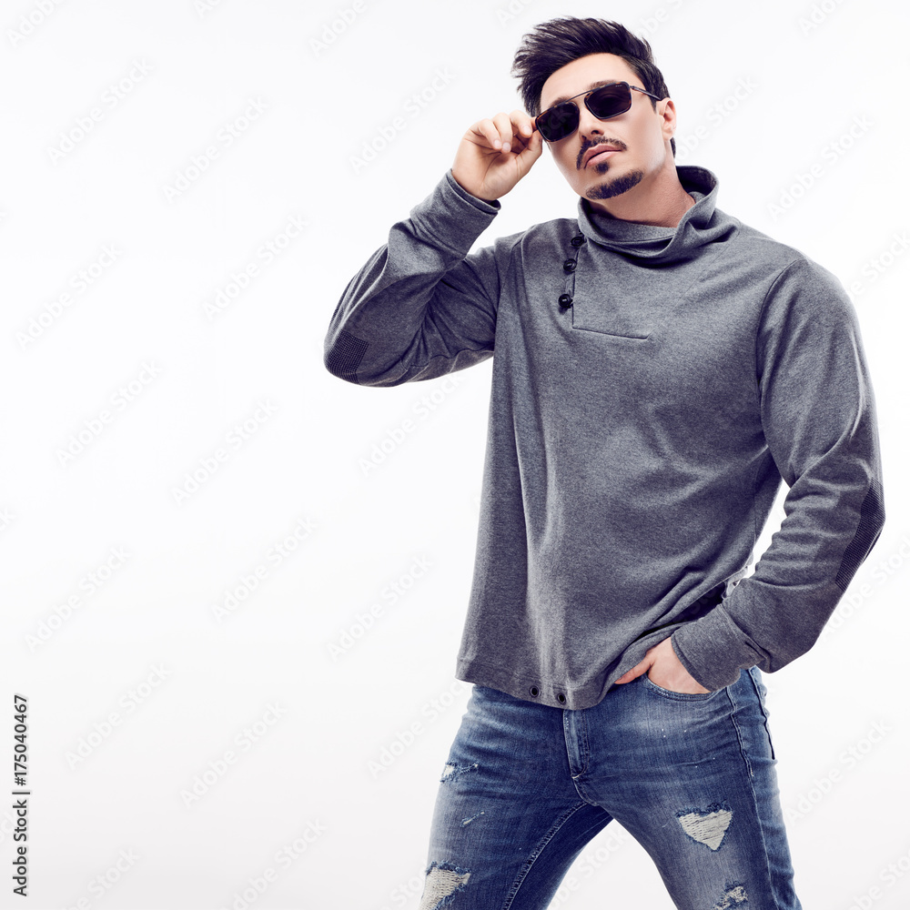 Handsome Sexy young Fashion guy in Trendy Sunglasses. Sporty Confident Brunette Bearded man. Autumn Outfit, Stylish Hairstyle, fashionable gray jumper. Studio fashion pose on white background Handsome Sexy young Fashion guy in Trendy Sunglasses. Sporty Confident Brunette Bearded man. Autumn Outfit, Stylish Hairstyle, fashionable gray jumper. Studio fashion pose on white background