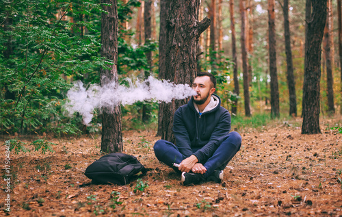 Vape man. brutal bearded young man having rest in forest. Man sitting under tree. Lifestyle. photo