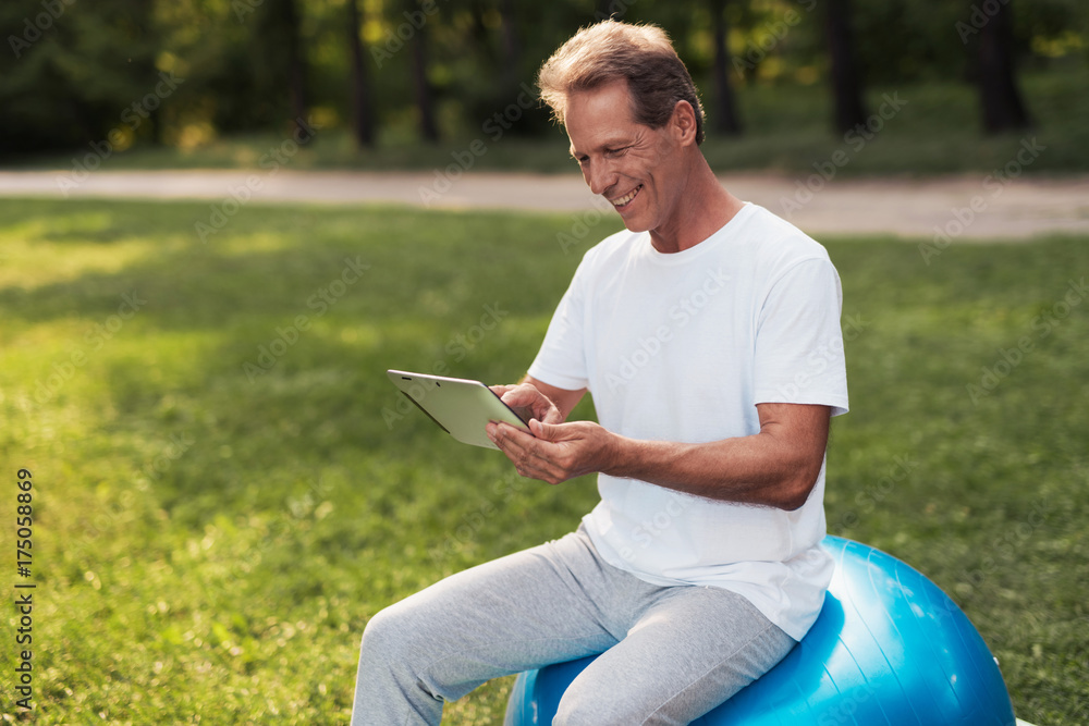 A man sits on a ball for yoga and looks at something on his tablet. He smiles
