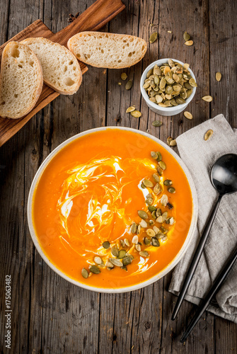 Traditional fall and winter dishes, hot and spicy pumpkin  soup with pumpkin seeds, cream and freshly baked baguette, on old rustic wooden table, copy space top view